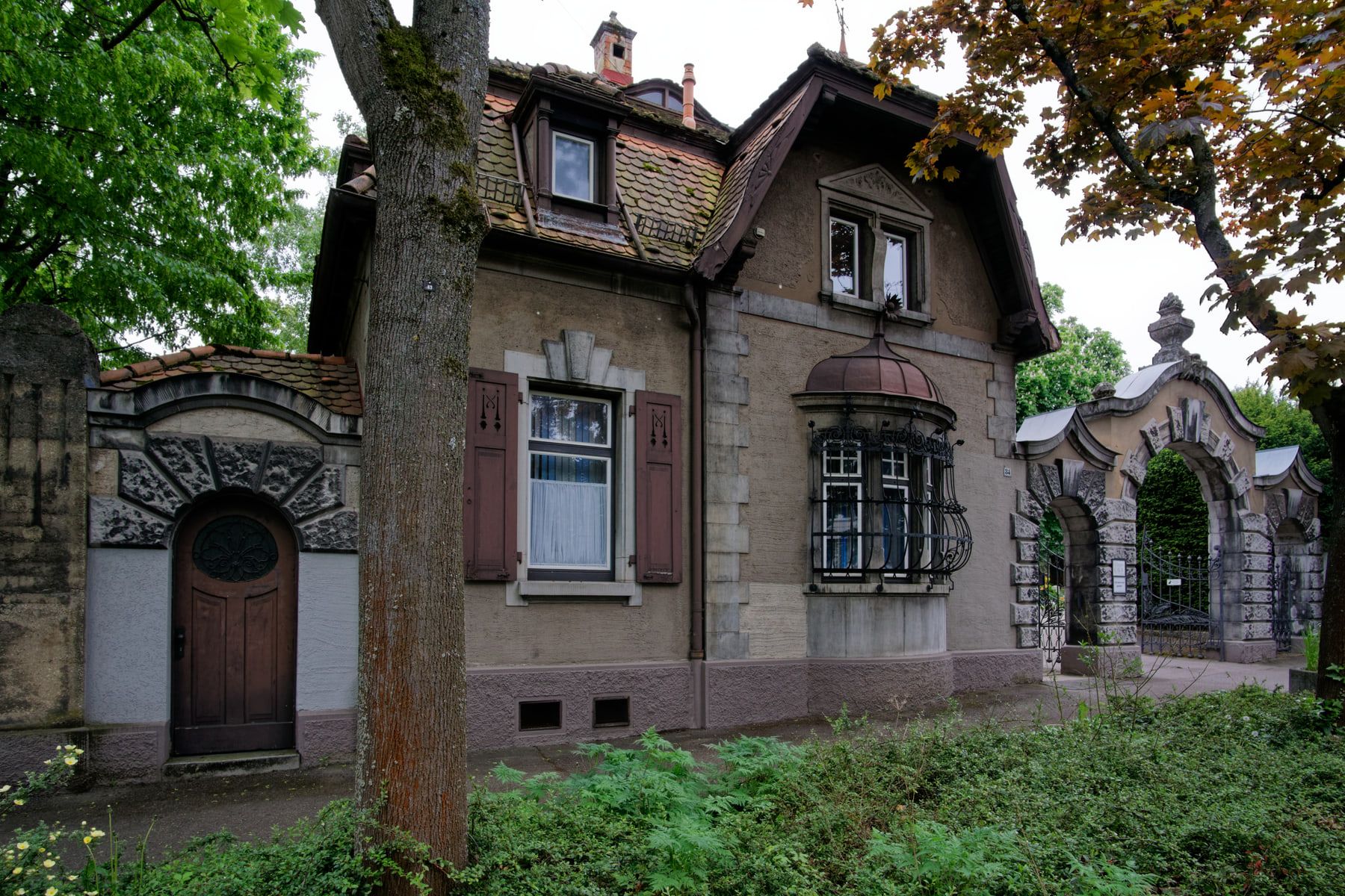Dienstgebäude am Portal mit Erker, Ziergitter und Ziegeldach zwischen alten Bäumen auf Bergfriedhof Stuttgart